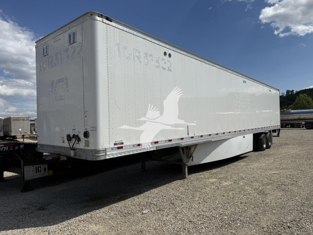 Trailer side view showing a white Hyundai Roll Door van trailer with a starling bird graphic and faded text along the side; dual rear axles near the back; gravel lot.
