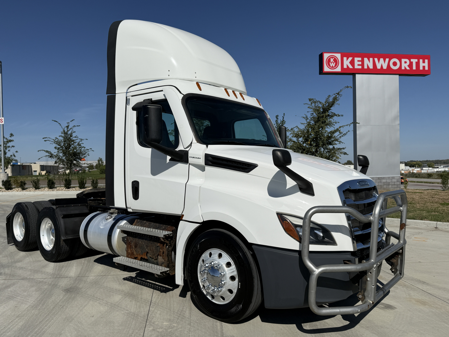 Truck cab exterior of a white 2021 Freightliner PT126064S T day cab providing a front three-quarter view with grille guard, Kenworth sign in background, side exhaust, dual rear axles.