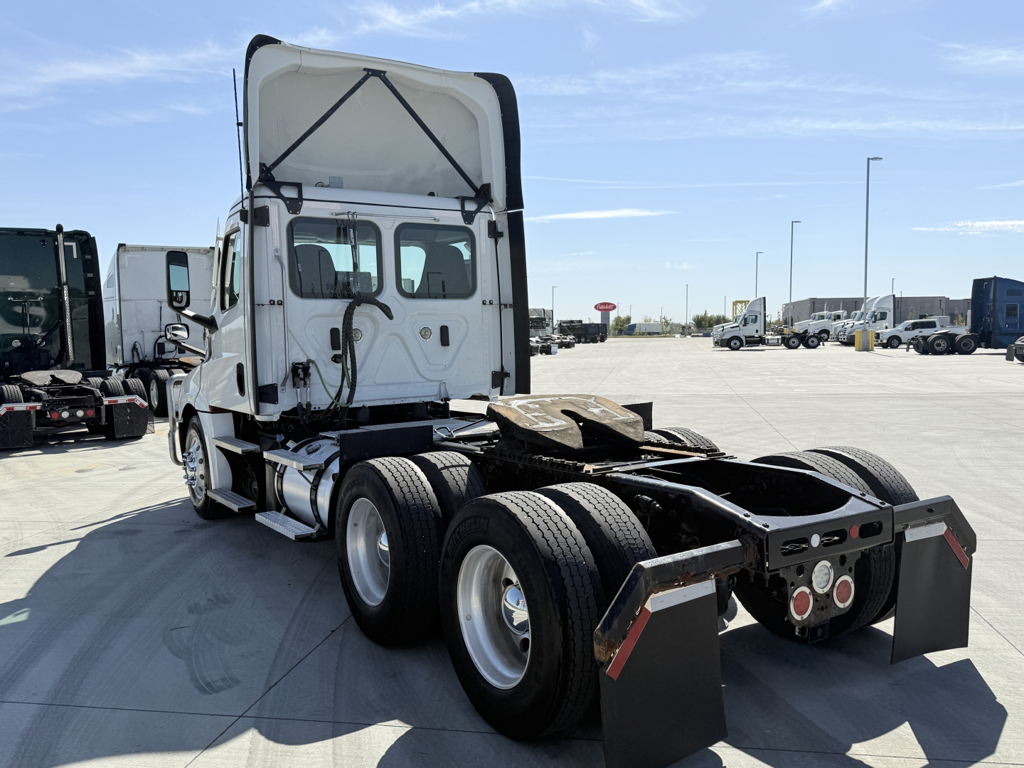 Truck shows rear view of a Freightliner daycab with tractor- trailer hitch, dual rear axles, and five-wheel mud flaps. Visible text on cab window area and chassis.