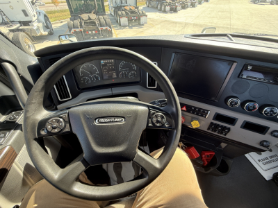 Steering wheel and dashboard inside a Freightliner PT126064S T cab. Visible dashboard gauges, steering wheel with Freightliner badge, center console with controls, and a red push-to-start/ignition area. The image shows a modern truck interi