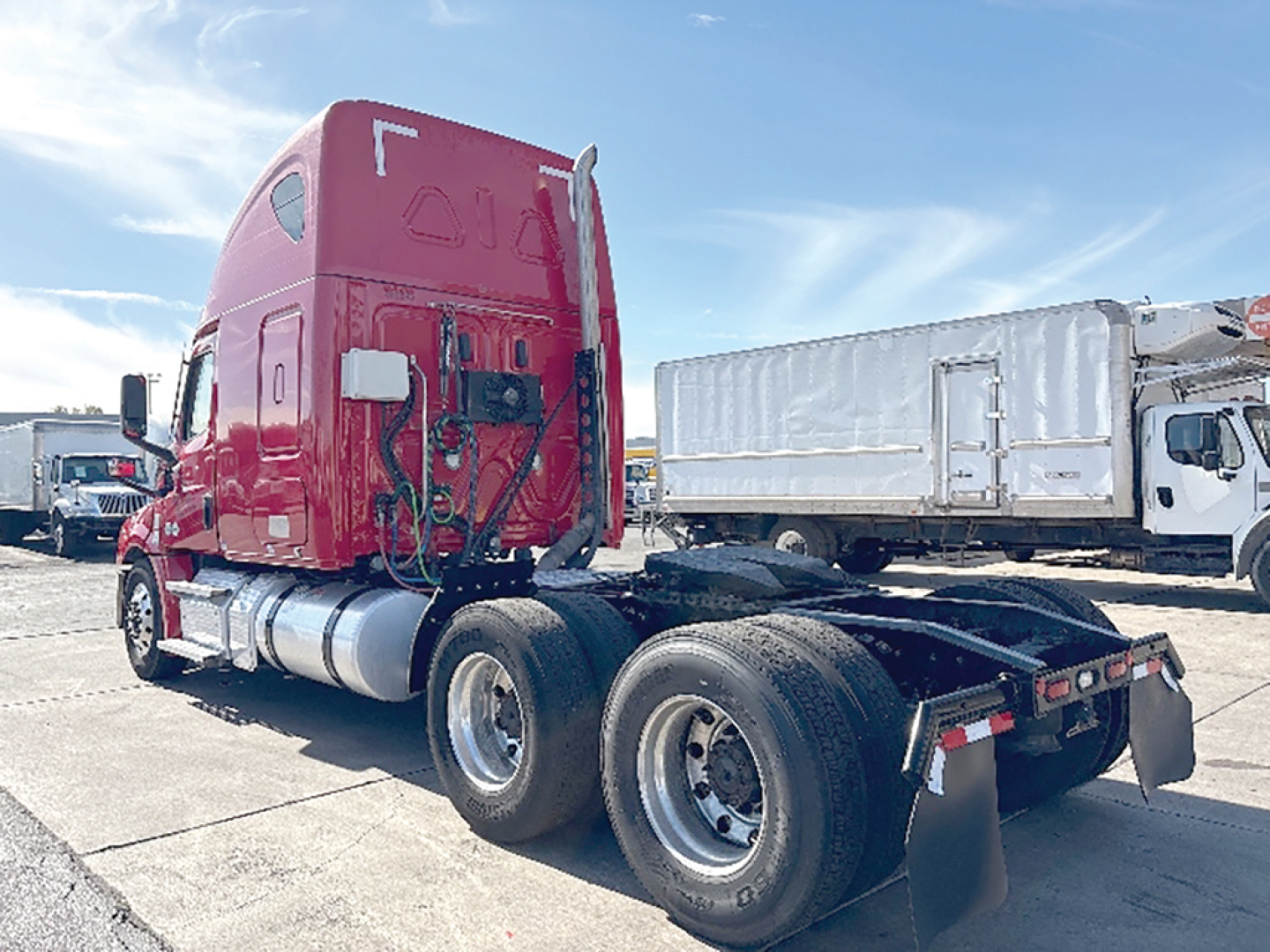 Red Freightliner Cascadia tractor with sleeper cab, rear view showing dual rear wheels and fifth wheel, with a white dump truck in background.