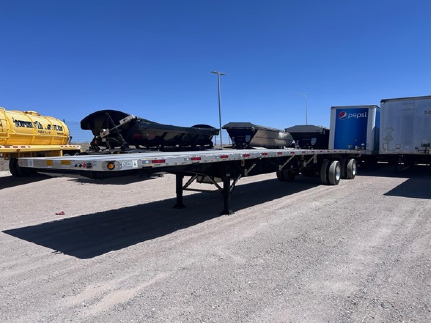 Flatbed trailer with a long low deck showing storage, with a nearby Pepsi trailer in a truck yard.