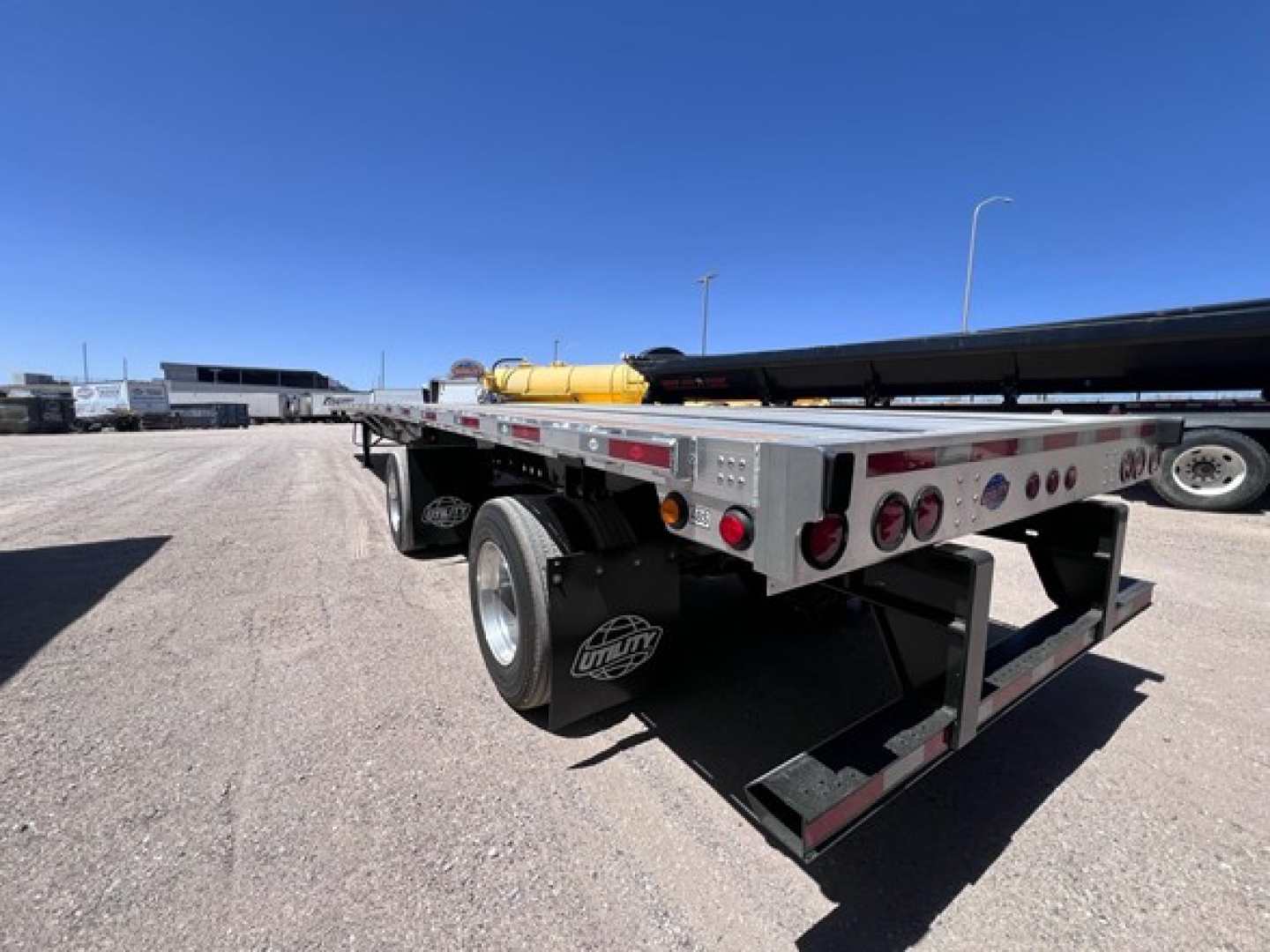 Flatbed trailer rear-left view showing dual rear axles and metal deck with yellow tank and equipment in background