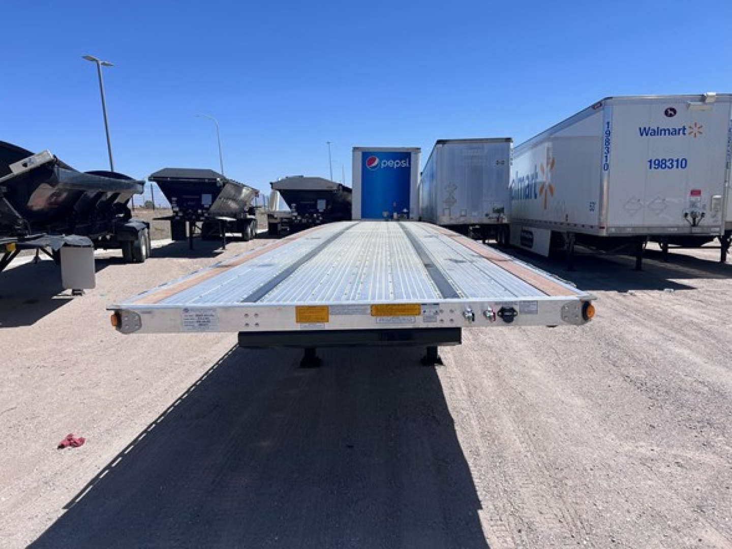 Flatbed trailer from rear view showing long silvery deck with safety lights and mounting hardware; trailers and trucks in background in a lot.