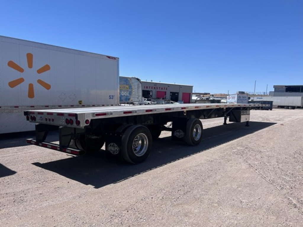 Trailer showing a long flatbed with dual rear wheels and a white deck in an outdoor lot.