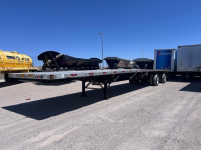 Flatbed trailer with a long low deck showing storage, with a nearby Pepsi trailer in a truck yard.