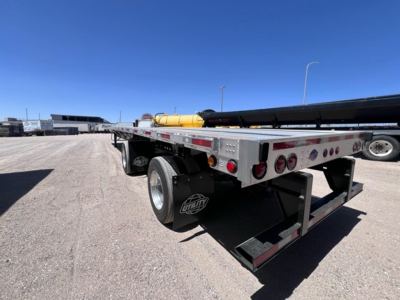 Flatbed trailer rear-left view showing dual rear axles and metal deck with yellow tank and equipment in background