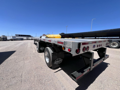 Flatbed trailer showing dual rear axles and metal frame with lights; spare wheel mudflap with 'US DOT' style sticker visible; yellow tank and equipment on site.