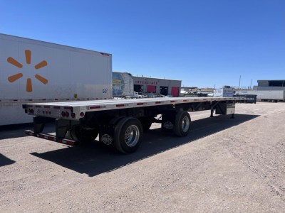 Long utility flatbed trailer in a yard with dual rear axles and a Walmart trailer in background.