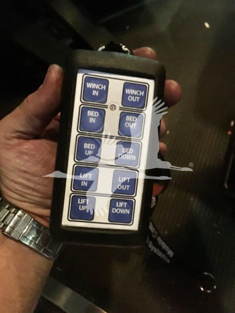 Remote control panel with blue labeled buttons for vehicle lift operations: WINCH IN, WINCH OUT, BED IN, BED OUT, BED UP, BED DOWN, LIFT IN, LIFT OUT, LIFT UP, LIFT DOWN.