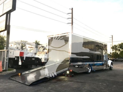 Truck bed with wheelchair-accessible ramp extended, connected to a large enclosed trailer on a rollback parking lot.