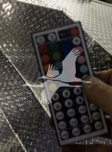 Remote control-like device with multicolor circular buttons being held over diamond plate metal floor; overlay logo obscures part of device.