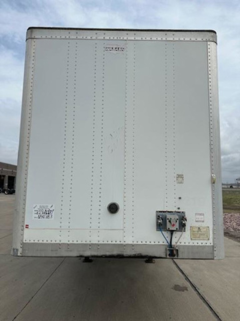 Rear view of a white Wabash trailer with riveted panels, doors closed, and a small control box near bottom right. Visible labels include a small sticker near bottom left and a circular door handle; thin vertical lines from rivets.