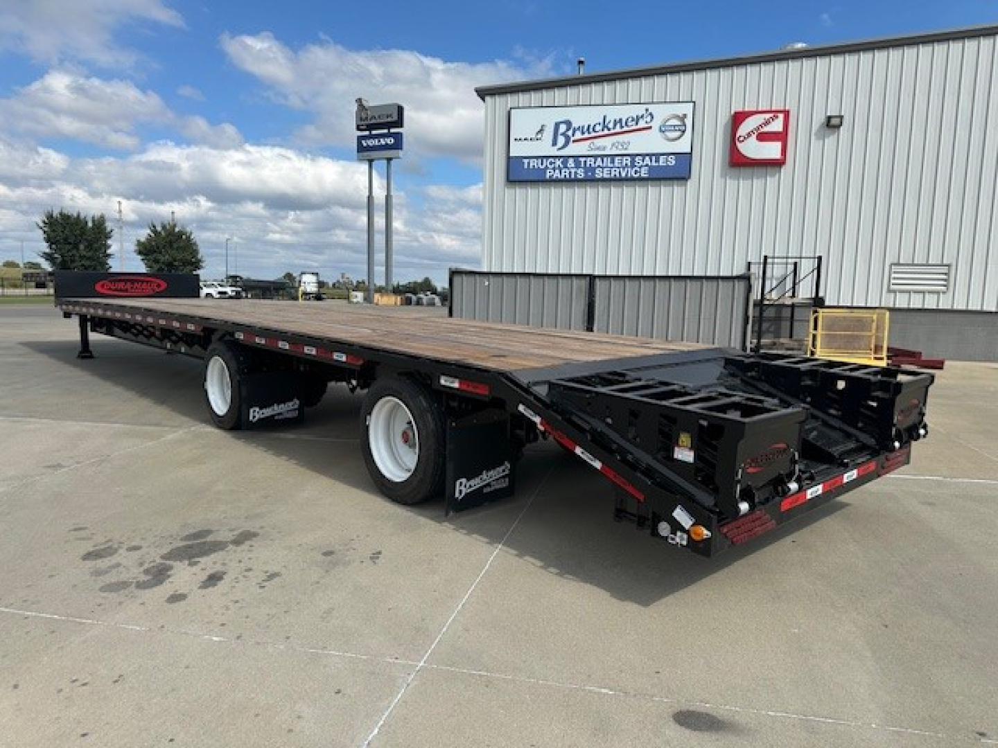 Trailer with long flatbed and drop deck, rear ramps extended, dual rear axles, heavy equipment trailer. Visible text: Ba... Buckner's? signage on building.