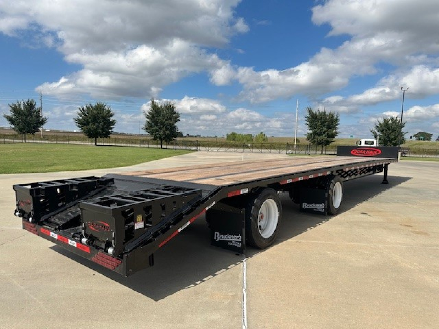 Trailer deck showing drop deck with tail ramps and spread configuration, black frame, wooden deck, three axles with white wheels.