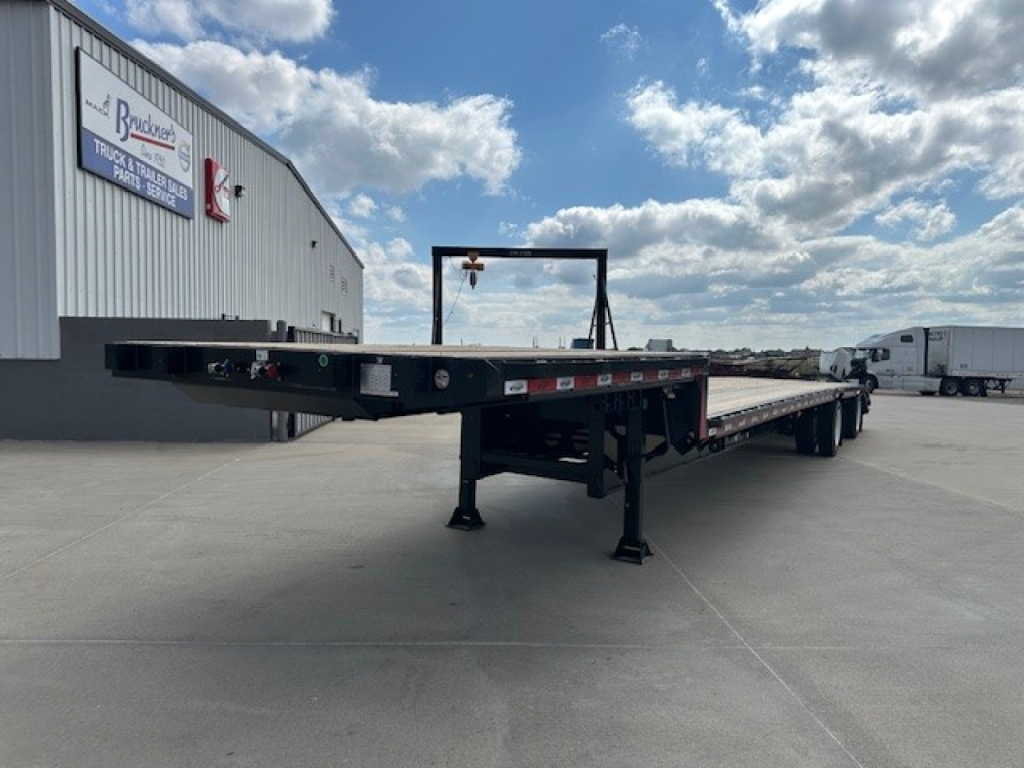 Trailer deck showing a long 53' drop deck trailer with spread tail ramps and support legs, at a dealership lot.