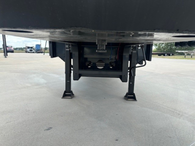 Drop deck trailer undercarriage with I-beam legs and ramp mechanism visible. No legible text or numbers in view.