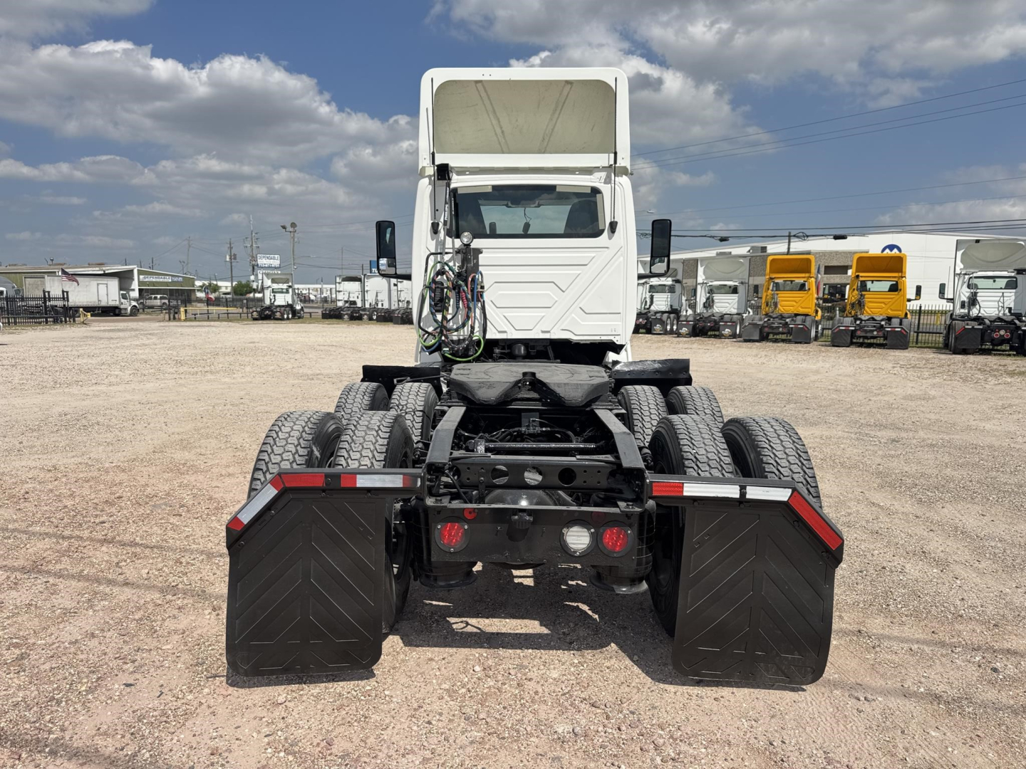 Tractor-trailer chassis rear view showing dual rear axles with large mud flaps and fifth-wheel area, white cab in distance, truck yard background.