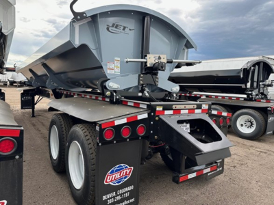 Jet brand side-dump trailer with a gray payload trough and rear dual wheels, showing the Jet logo, top view of dump body, coupling mechanism, and utility trailer wheels.
