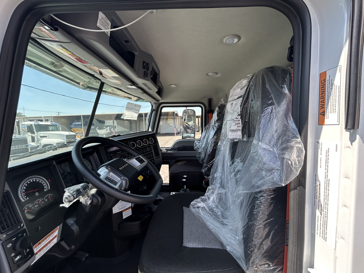 Interior cab of a new 2026 Mack GR64B rolloff truck showing steering wheel, dashboard, driver seat covered in plastic, and passenger seat; VIN and stock tag not visible in image.