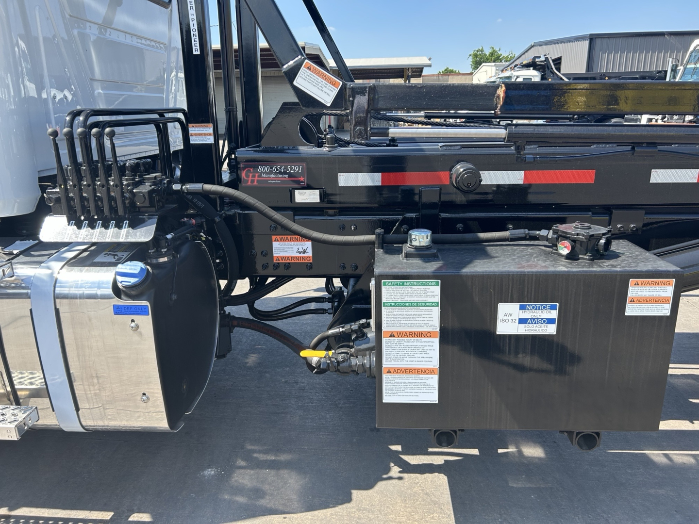 Truck side view showing control box and hydraulic system on Mack GR64B roll-off truck with labels and warning stickers.