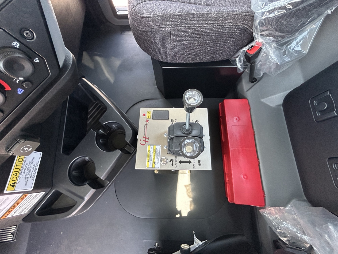 Minimum view of a Mack GR64B truck cab interior showing gear selector with two levers on a silver control plate, black steering area, red item storage, and chair with plastic cover.