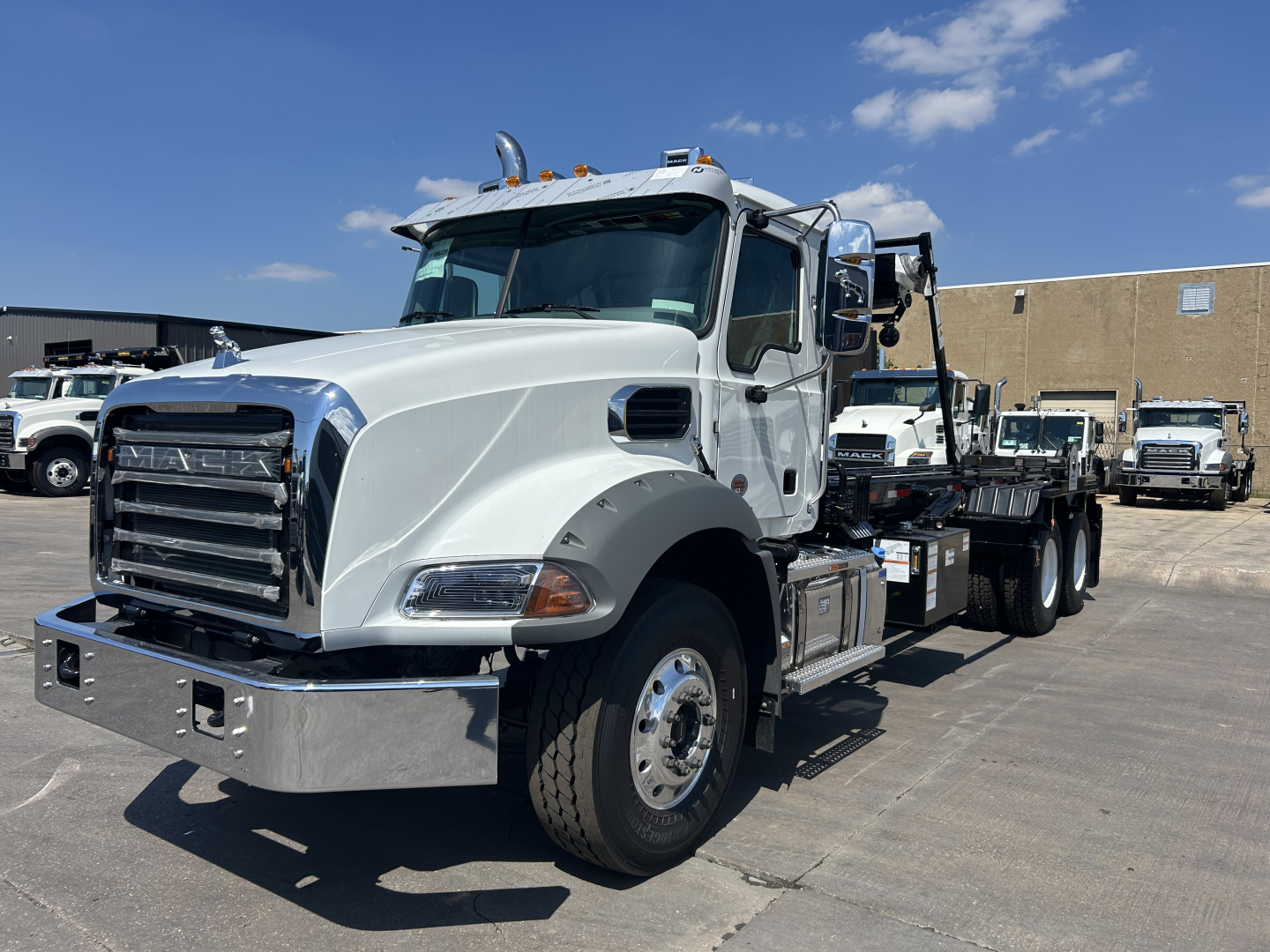 Truck: 2026 Mack GR64B roll-off chassis, white cab with chrome front bumper, dual rear axles, flatbed crane apparatus, VIN plate and dealership lot in background.