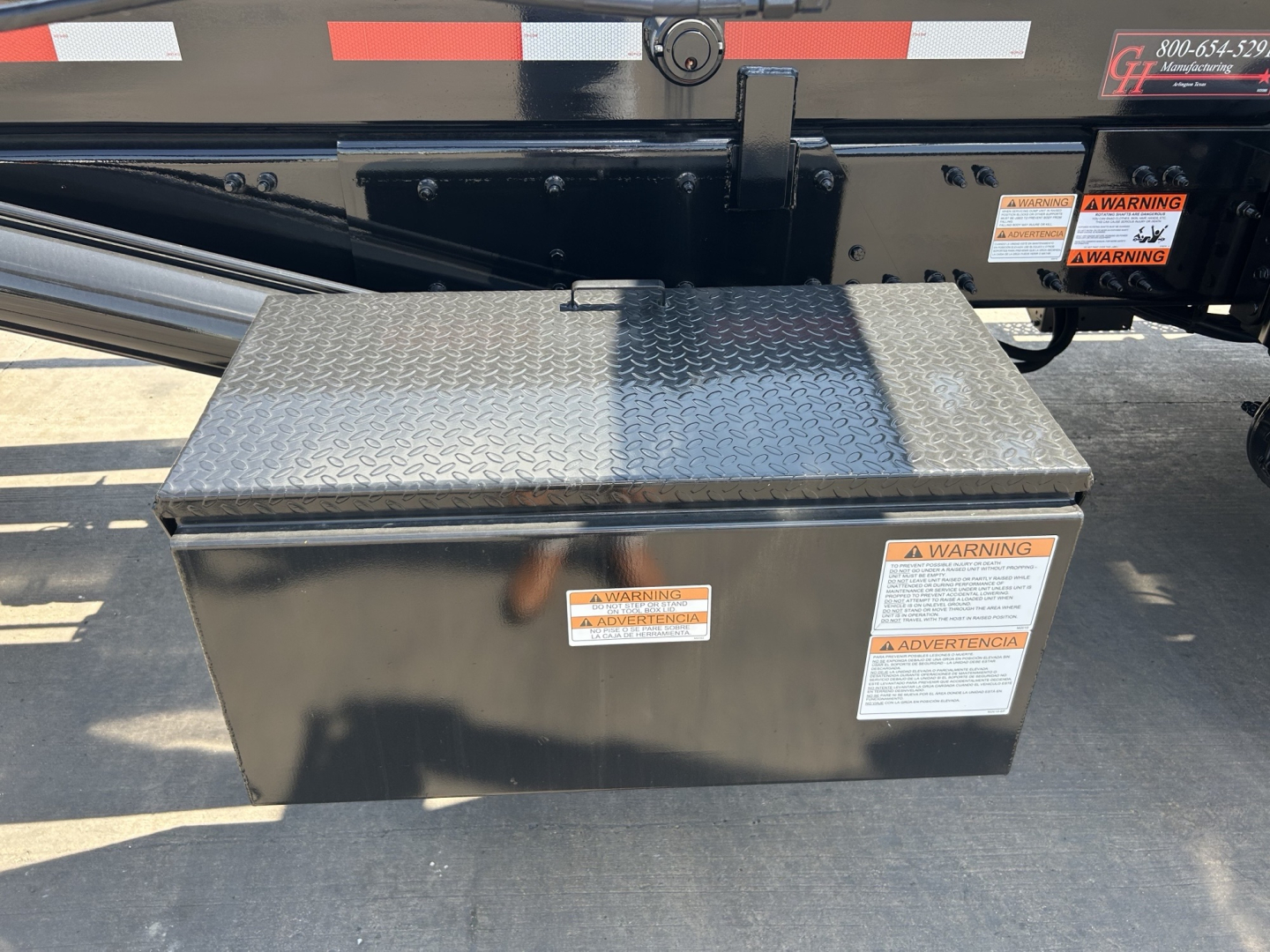 Rolloff truck hitch area with diamond-plate toolbox mounted on side of chassis; caution labels on toolbox and side rail; black painted metal frame with reflective stripes.