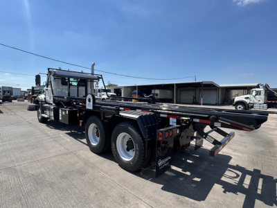 Truck bed of a new 2026 Mack GR64B roll-off truck, side view showing long flatbed and dual rear axles with heavy-duty tires and rear ramps.