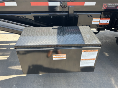 Rolloff truck hitch area with diamond-plate toolbox mounted on side of chassis; caution labels on toolbox and side rail; black painted metal frame with reflective stripes.