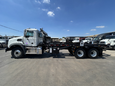 2026 Mack GR64B rolloff truck side view showing Mack cab, chassis, and multiple axles with flatbed. Visible text on components includes no legible labels from distance.