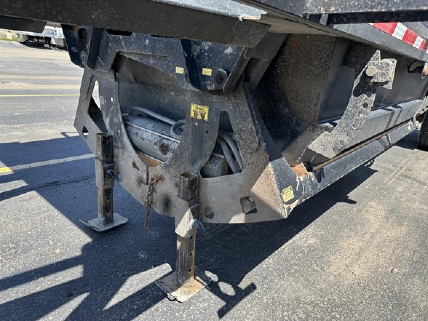 Suspension and undercarriage of a bottom dump trailer with hydraulic components and frame hardware. Visible labels and yellow warning stickers on metal brackets.