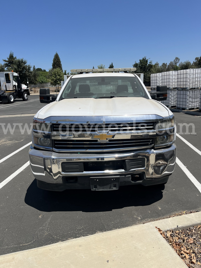 Chevrolet Silverado 2500HD truck front view with chrome grille and bumper, Chevrolet emblem, dealer watermark across windshield, and flatbed/dump setup in a parking lot.