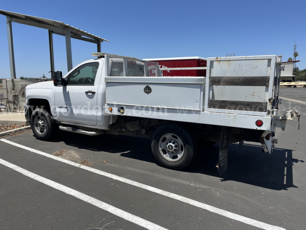 Flatbed dump truck side view showing white 2015 Chevrolet Silverado 2500HD with metal utility bed and diamond plate storage boxes.
