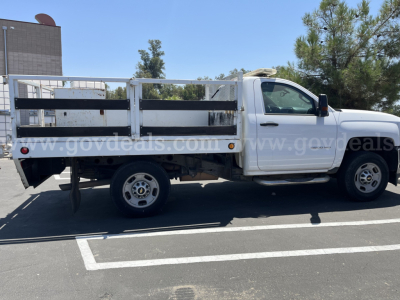 Flatbed dump truck side view showing white 2015 Chevrolet Silverado 2500HD with rectangular steel stake bed and black side rails.