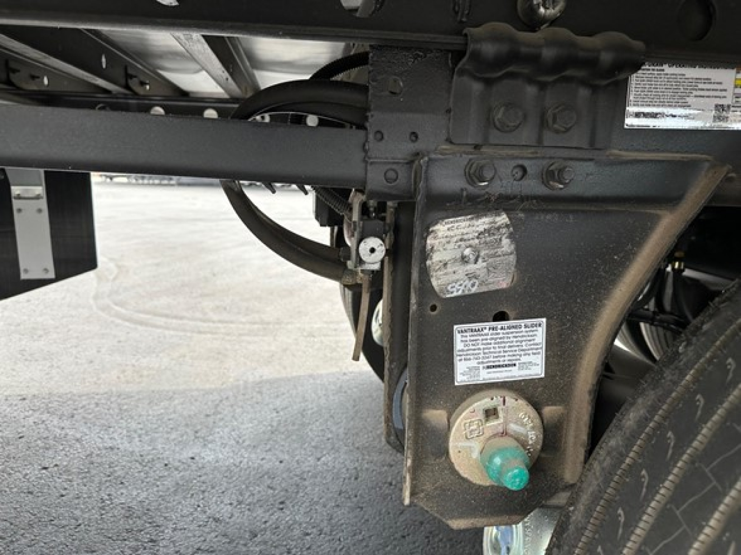 Trailer frame with label and valve showing various stickers and a green capped fitting; close-up of chassis area under a reefer trailer with riveted plate and wiring.