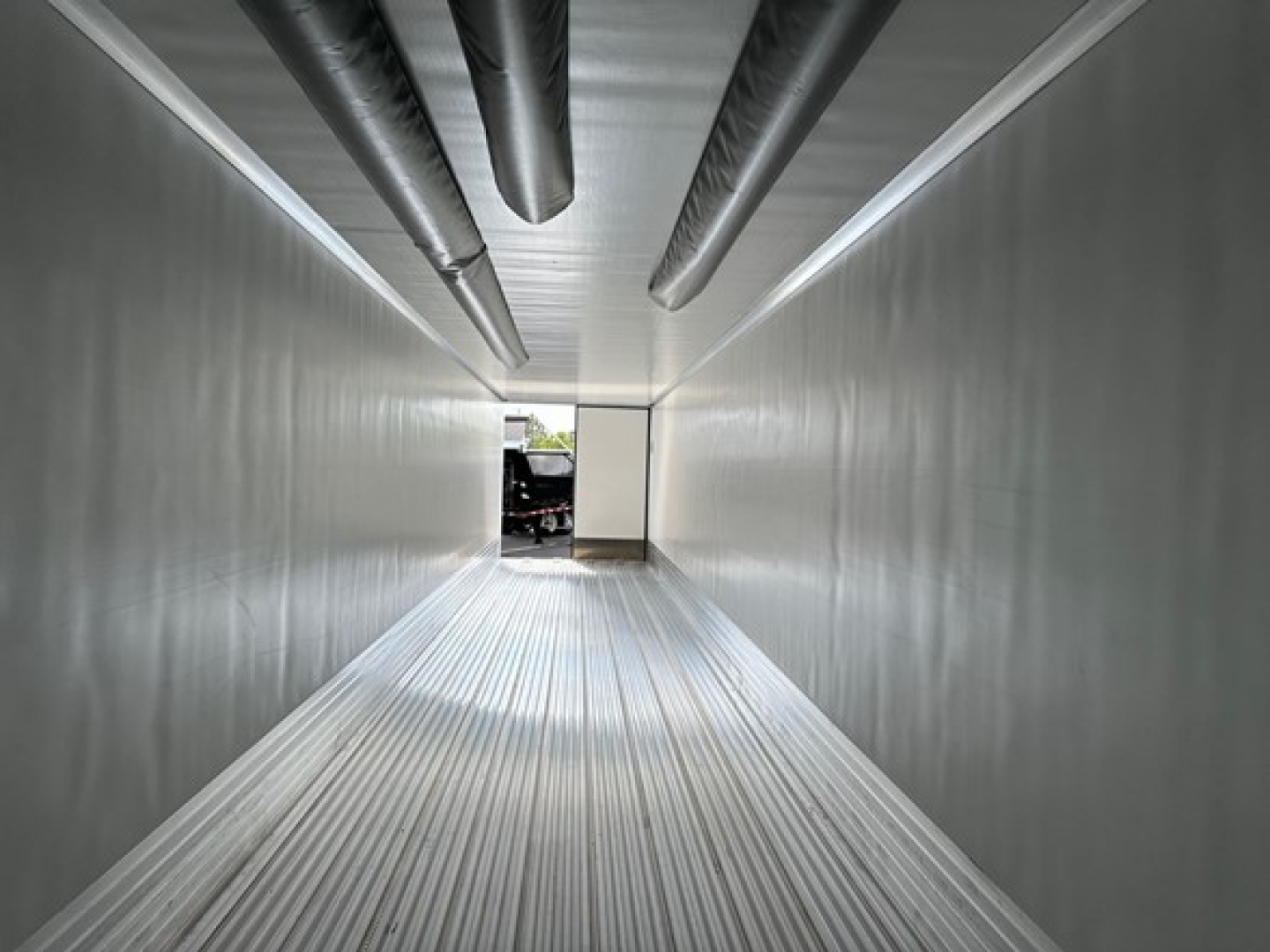 Trailer interior showing metal walls and ceiling with ducts, long narrow corridor, light at far end.