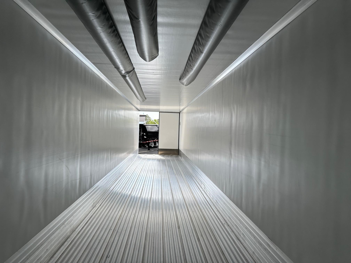 Trailer interior showing metal walls and ceiling with hanging pipes, long aluminum floor, view toward rear opening.