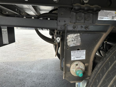 Trailer support frame with hoses and a circular green cap, label stickers, and a rectangular metal plate near the wheel well; close-up view of rear undercarriage of a trailer.
