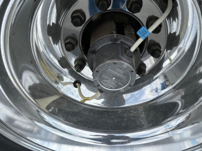 Trailer wheel hub with chrome rim showing a central hub cap, hydraulic line with blue tag, and polished surface reflecting surroundings.