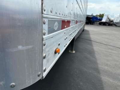 Trailer side view showing riveted aluminum wall with orange side marker, reflective stripe, and support leg on a blacktop lot.