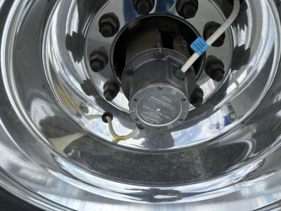 Wheel hub with chrome rim showing tire valve, brake drum and a label on a blue zip-tie. Visible text on the center cap reads 'SKF' and model numbers.