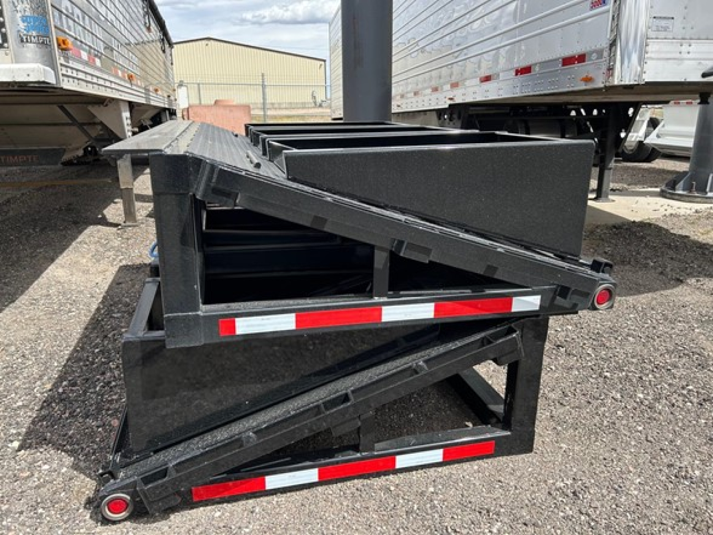 Trailer accessory showing folded beaver tail ramps and frame with reflective red/white tape. View from side with other trailers in background.