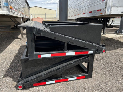 Trailer accessory showing folded beaver tail ramps and frame with reflective red/white tape. View from side with other trailers in background.