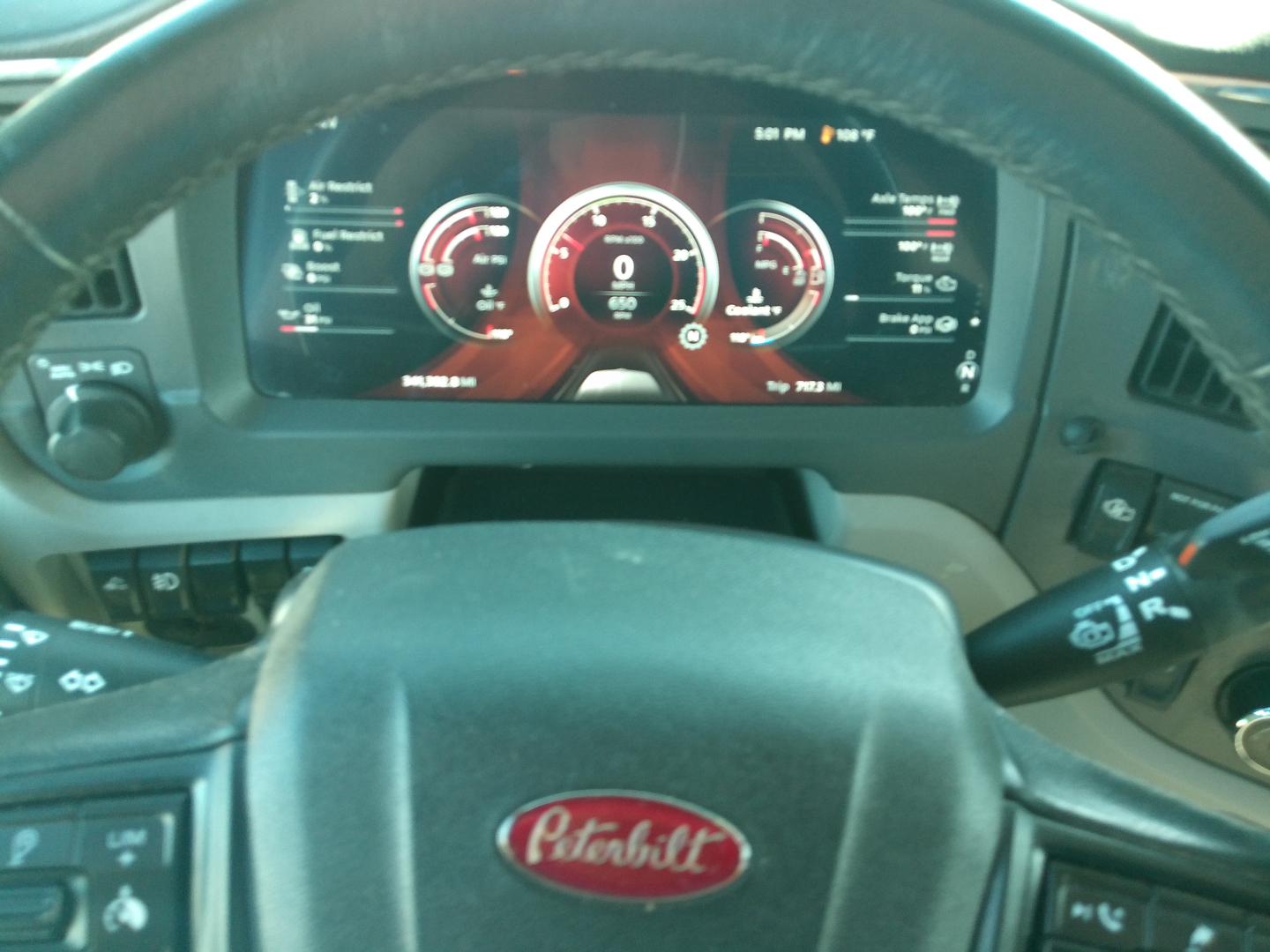Peterbilt dashboard view from steering wheel showing digital gauges in red; Peterbilt emblem on steering wheel; faint left and right stalks; center digital display.