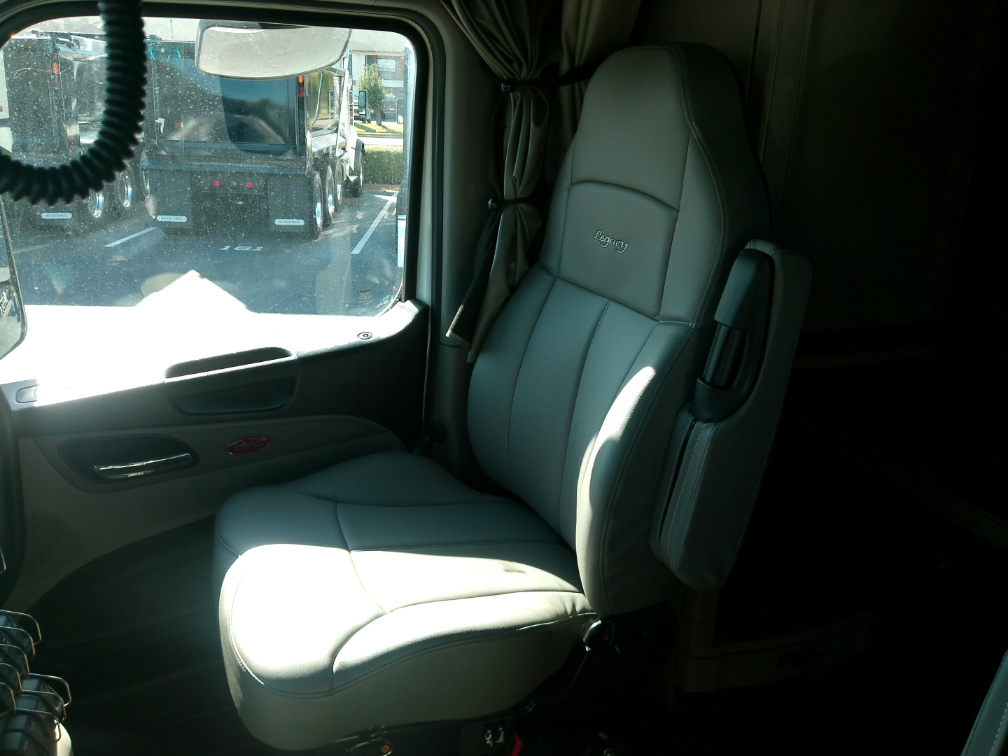 Truck cab interior showing grey driver seat and door panel in a Peterbilt truck with window view of another truck outside.