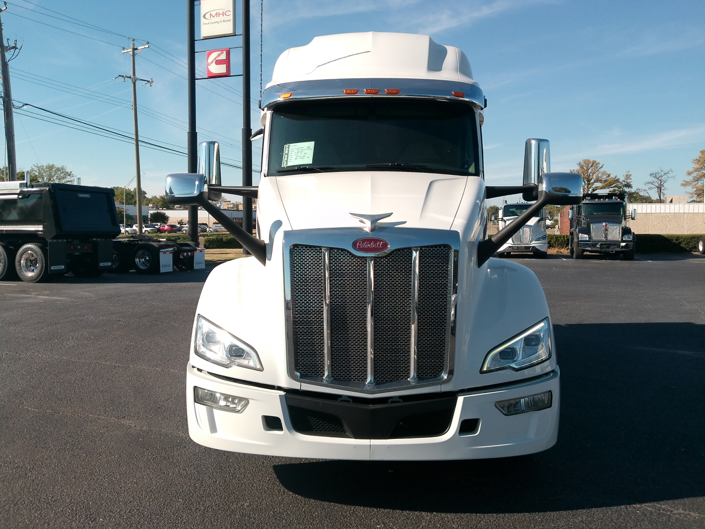 Peterbilt 579 truck front view in white with large grille, side mirrors, and tall cab, parking lot with other trucks in background.