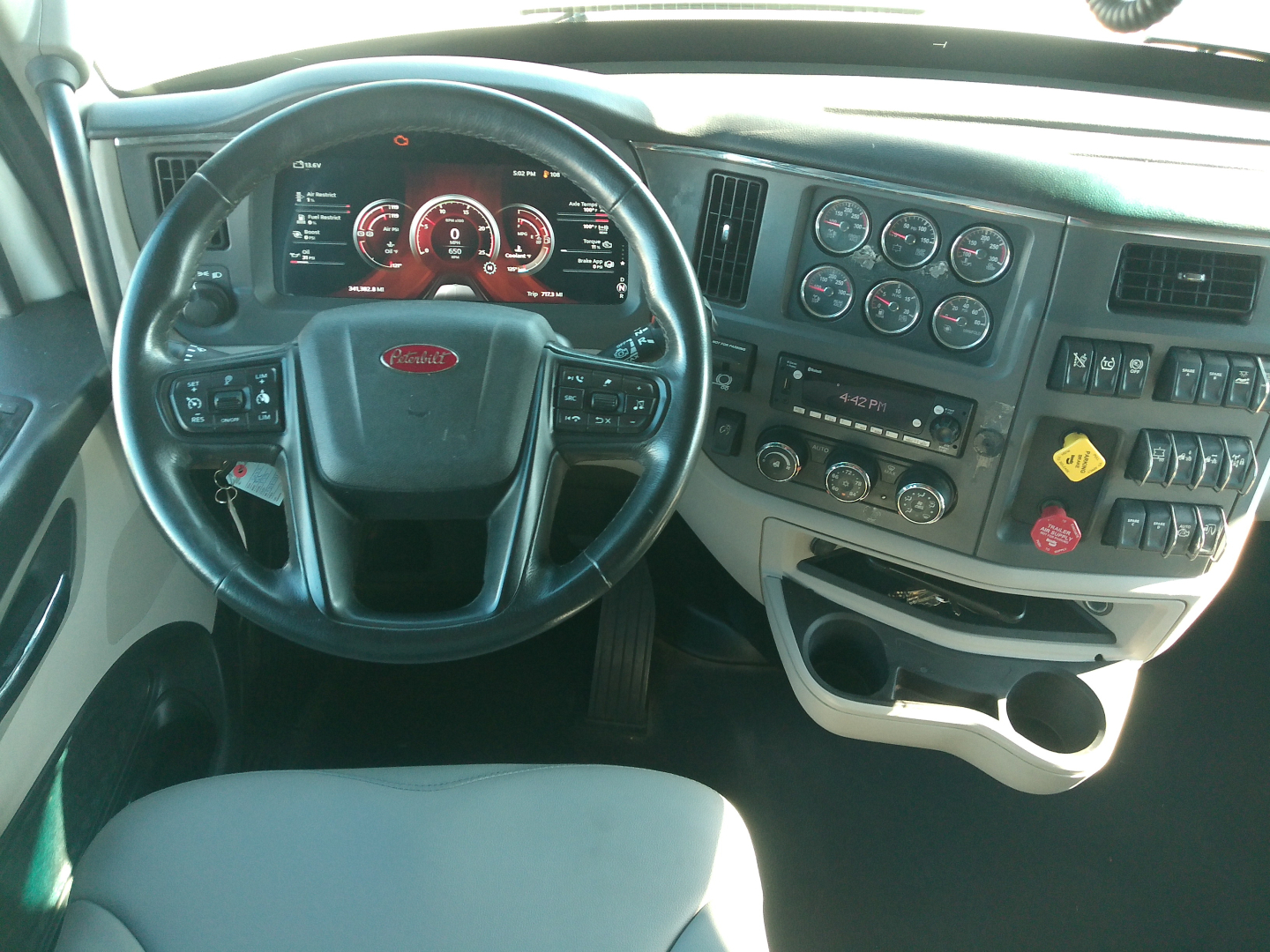 Peterbilt truck interior showing steering wheel with Peterbilt emblem and instrument cluster, dashboard controls, gauges, and beige lower panel.