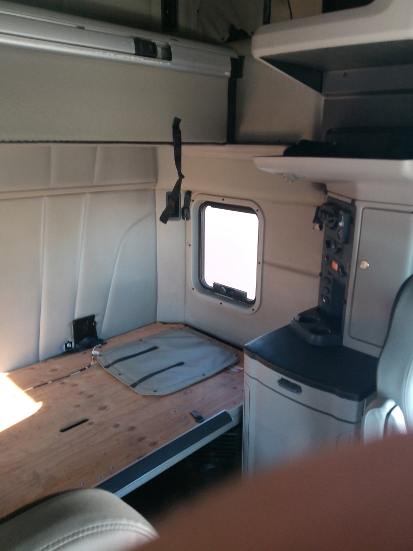 Peterbilt 579 truck cabin interior showing sleeper area with bed platform, small side window, and storage cabinet; light beige/gray interior.