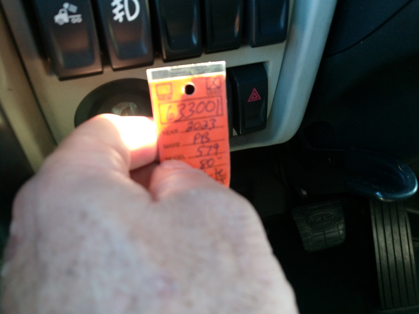 Tag being held in truck interior with dashboard controls and pedals in view; handwritten tag with numbers and letters.